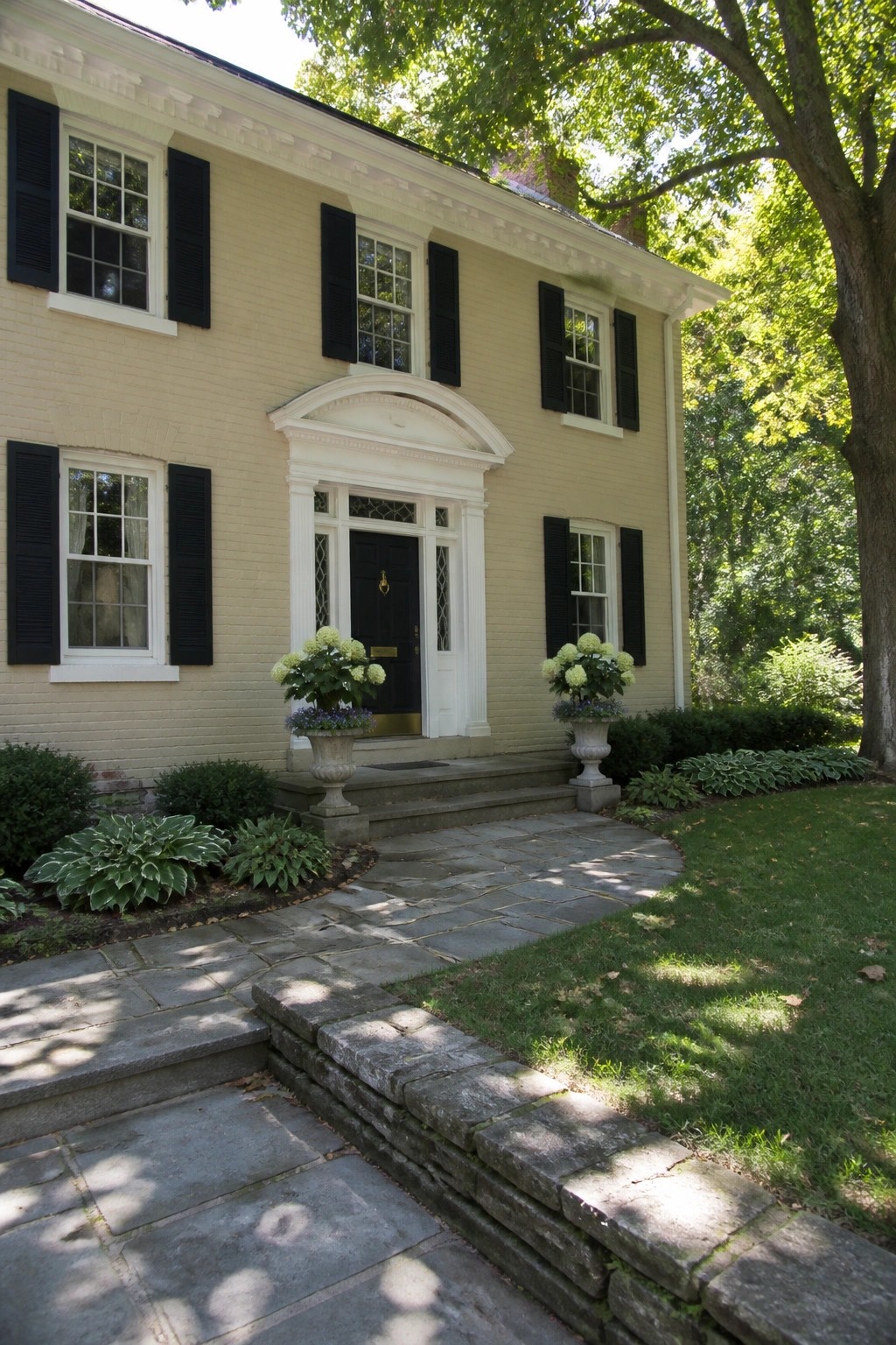 Yellow brick house with black shutters, white portico, and urns with white flowers