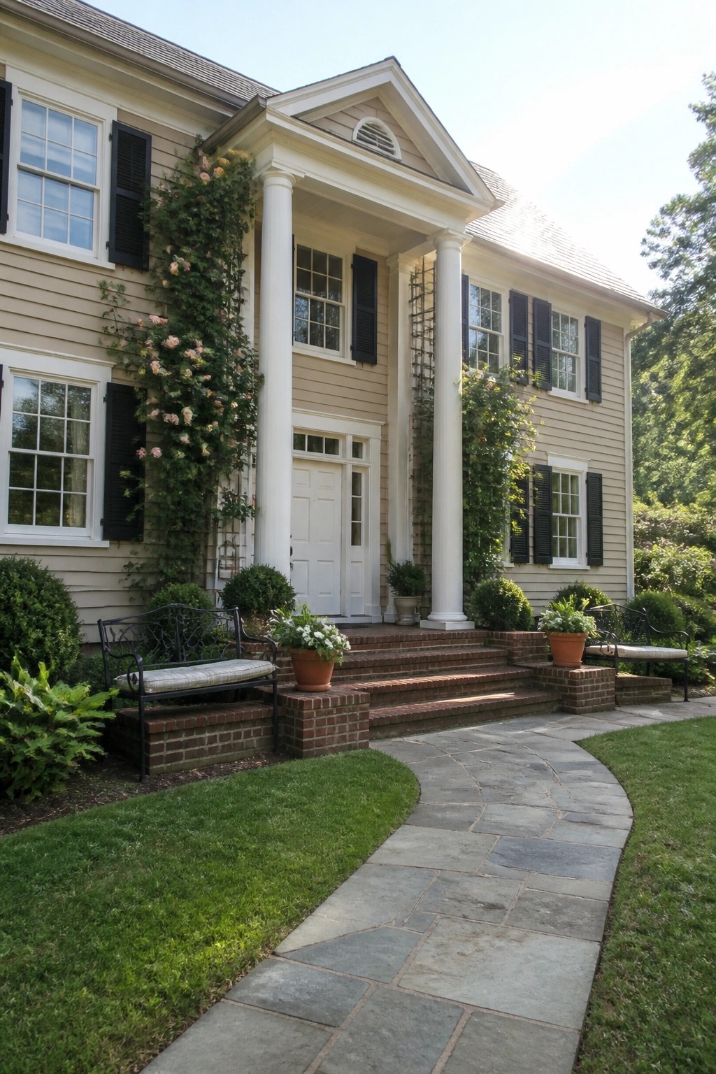 A beige colonial house with white columns, trim, and black shutters, featuring a brick walkway and climbing roses on the left side.