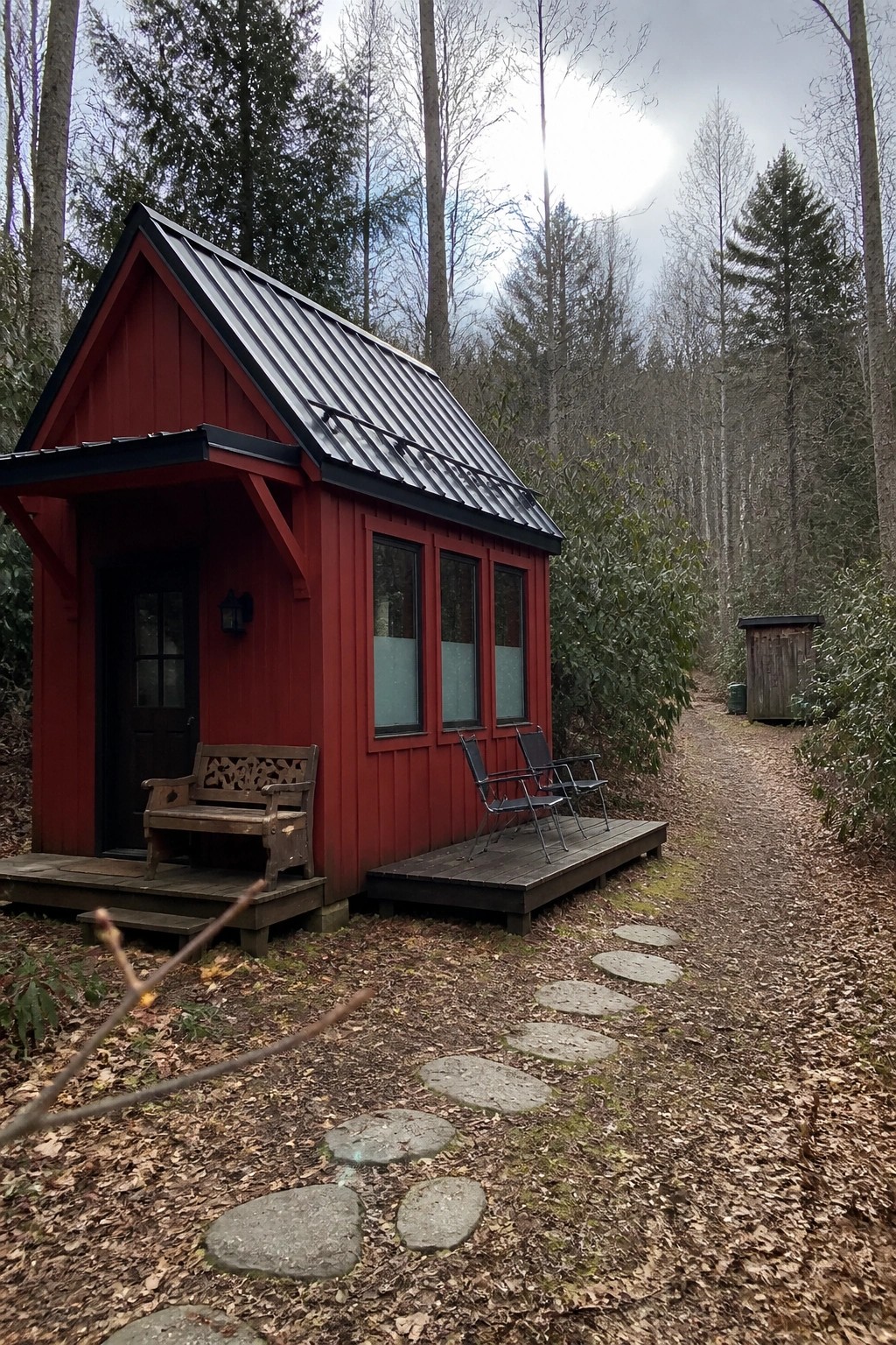 A small red cabin with black trim, a metal roof, and a wooden deck sits among trees with stepping stones leading to the front.