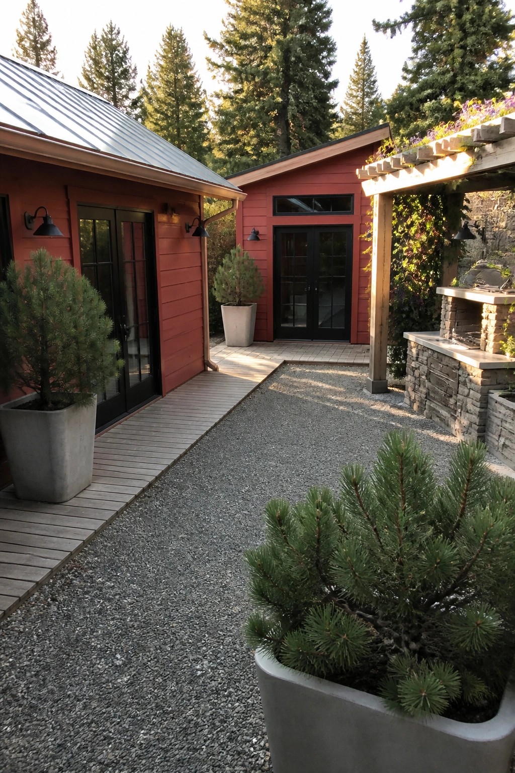 A red wooden cabin exterior with black-framed doors, a wooden deck, gravel path, and potted evergreens next to a stone outdoor kitchen under a pergola.