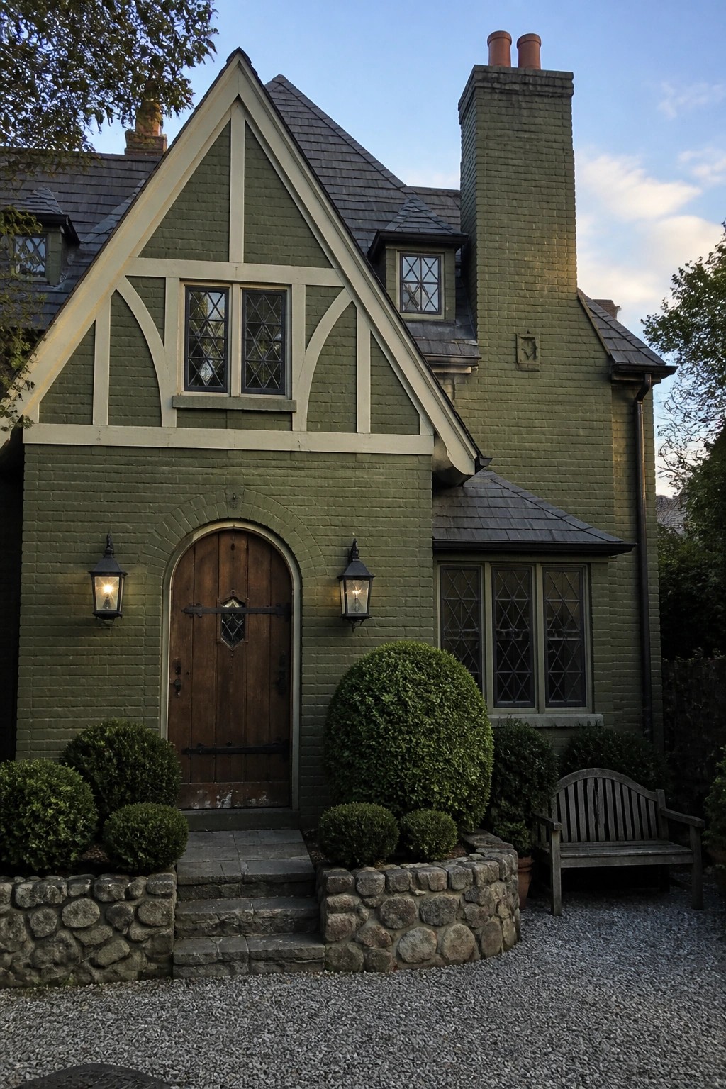 Green brick Tudor house with arched door, diamond windows, shrubs, and bench.