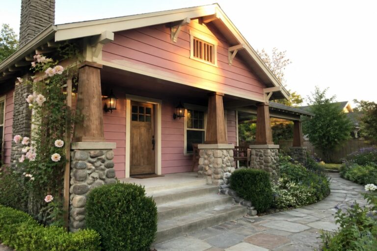 Pink cottage with stone pillars, wooden porch, steps, and blooming garden path.