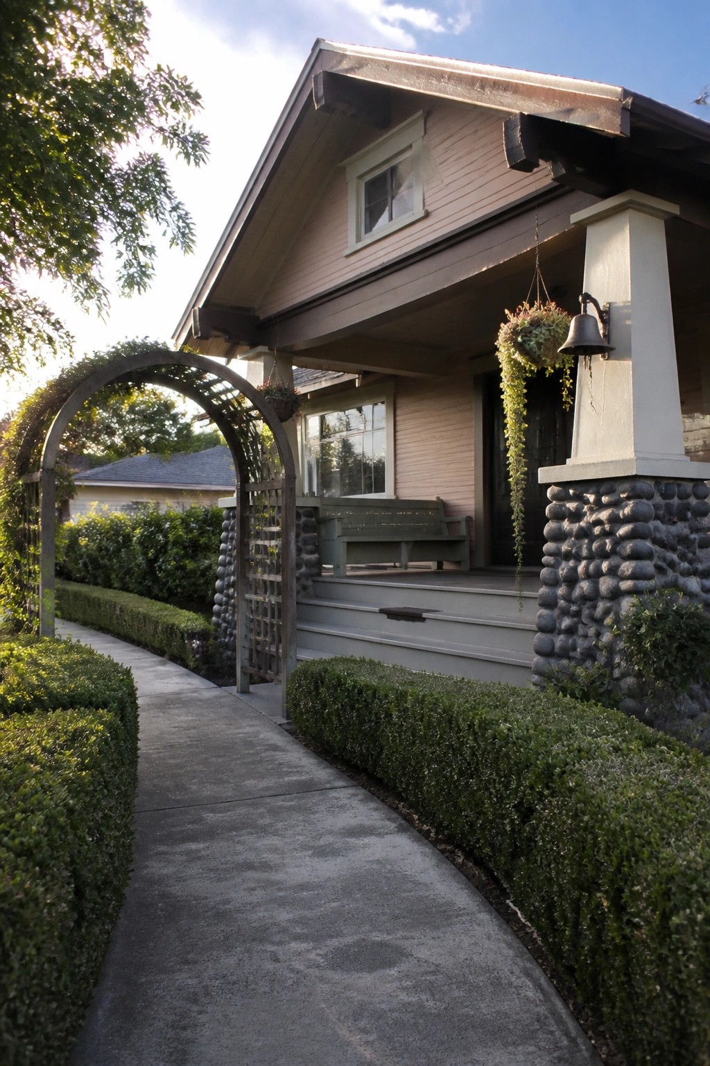 A pink Craftsman house exterior with a stone pillar supporting the porch, a wooden garden arch over a concrete path, and trimmed hedges along the walkway.