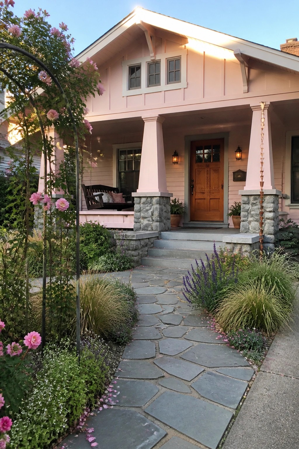 A pink Craftsman house with a wooden front door, stone porch columns, gray stone steps, and a stone pathway bordered by pink roses and purple flowers.