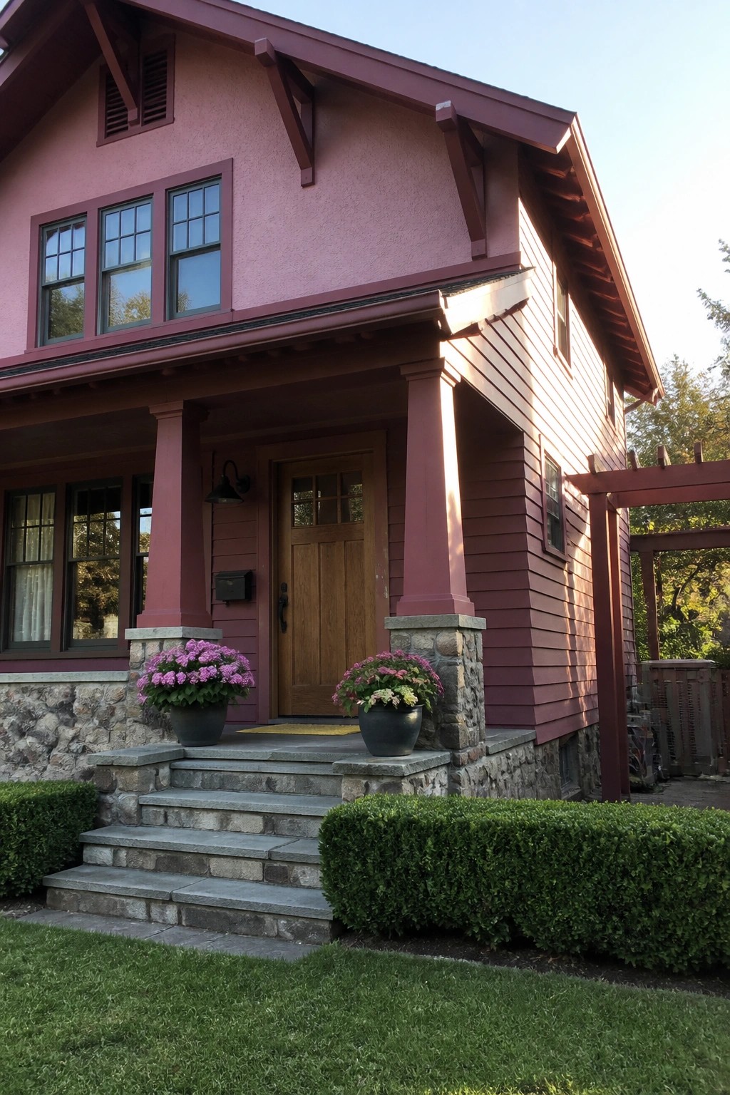A pink Craftsman house exterior showing a front porch with red wooden columns resting on stone bases, a wooden door, and stone steps with potted plants.