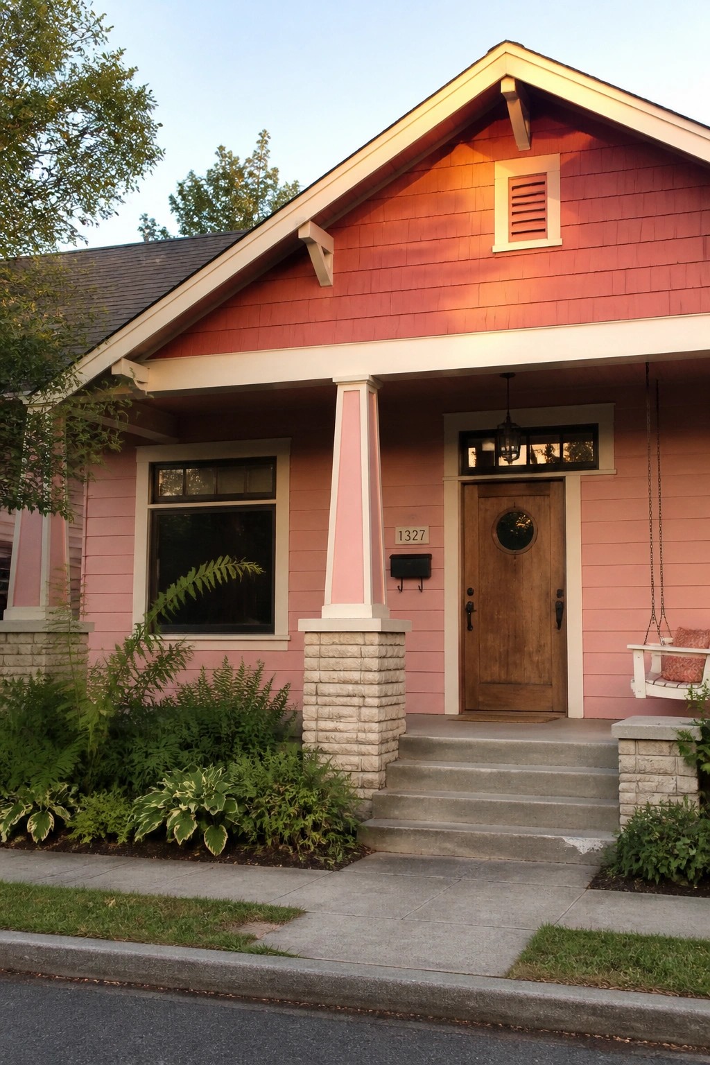 A pink Craftsman house with white trim, stone porch columns, a wooden front door, and concrete steps leading to the entry.