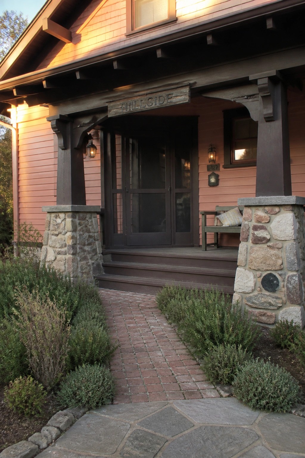 Front view of a pink Craftsman house with dark wood trim, stone porch pillars, wooden door, and brick path leading to the steps.