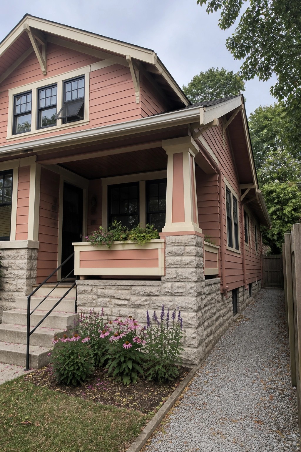 A pink Craftsman house exterior showing wood siding, stone foundation and columns, a covered porch, and flower beds along the front.