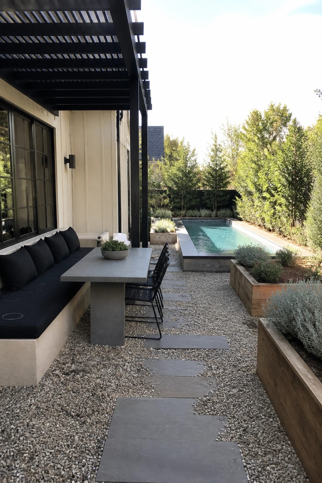 A covered outdoor patio features a long concrete dining table with a built-in bench holding black cushions, several black metal chairs, a black pergola overhead, and a rectangular pool visible beyond gravel paths and wooden planters.