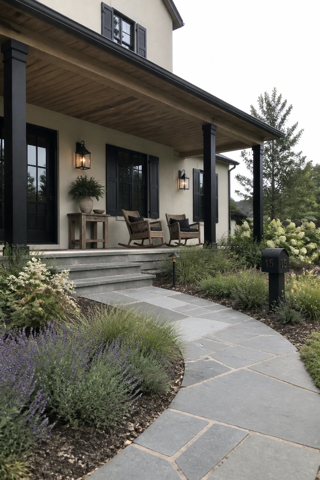 A beige house exterior with black columns, shutters, and door, featuring a covered porch with two rocking chairs, stone steps, and a curved stone walkway leading to a black mailbox labeled 15.