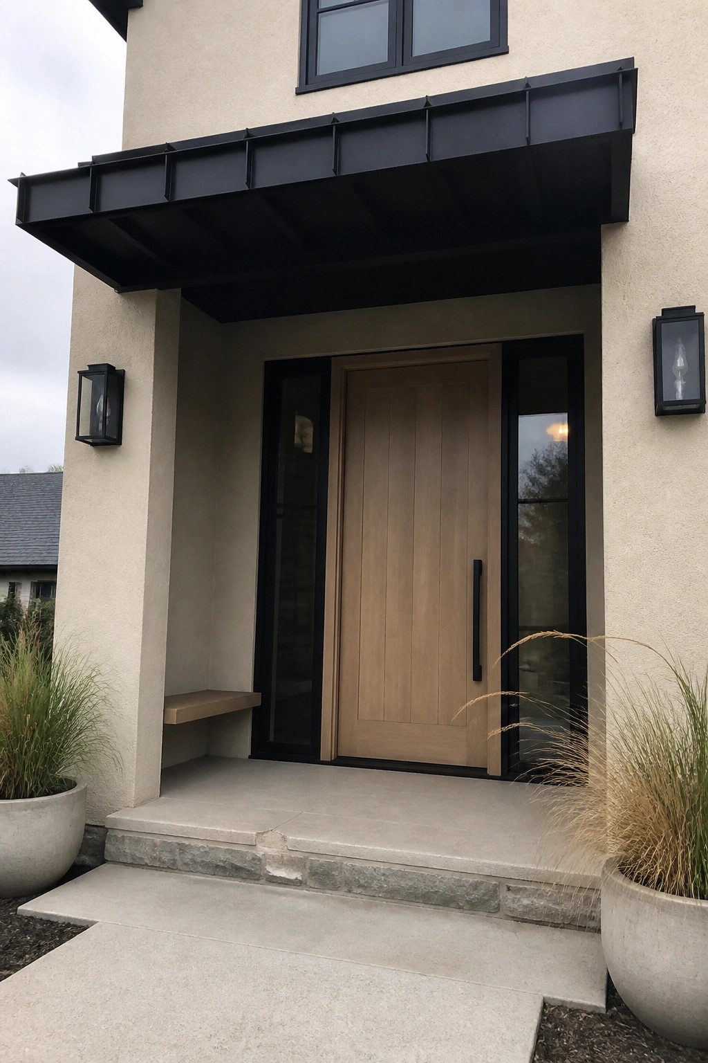 A beige stucco house exterior with a black metal awning over a light wood front door and black-framed glass sidelights.