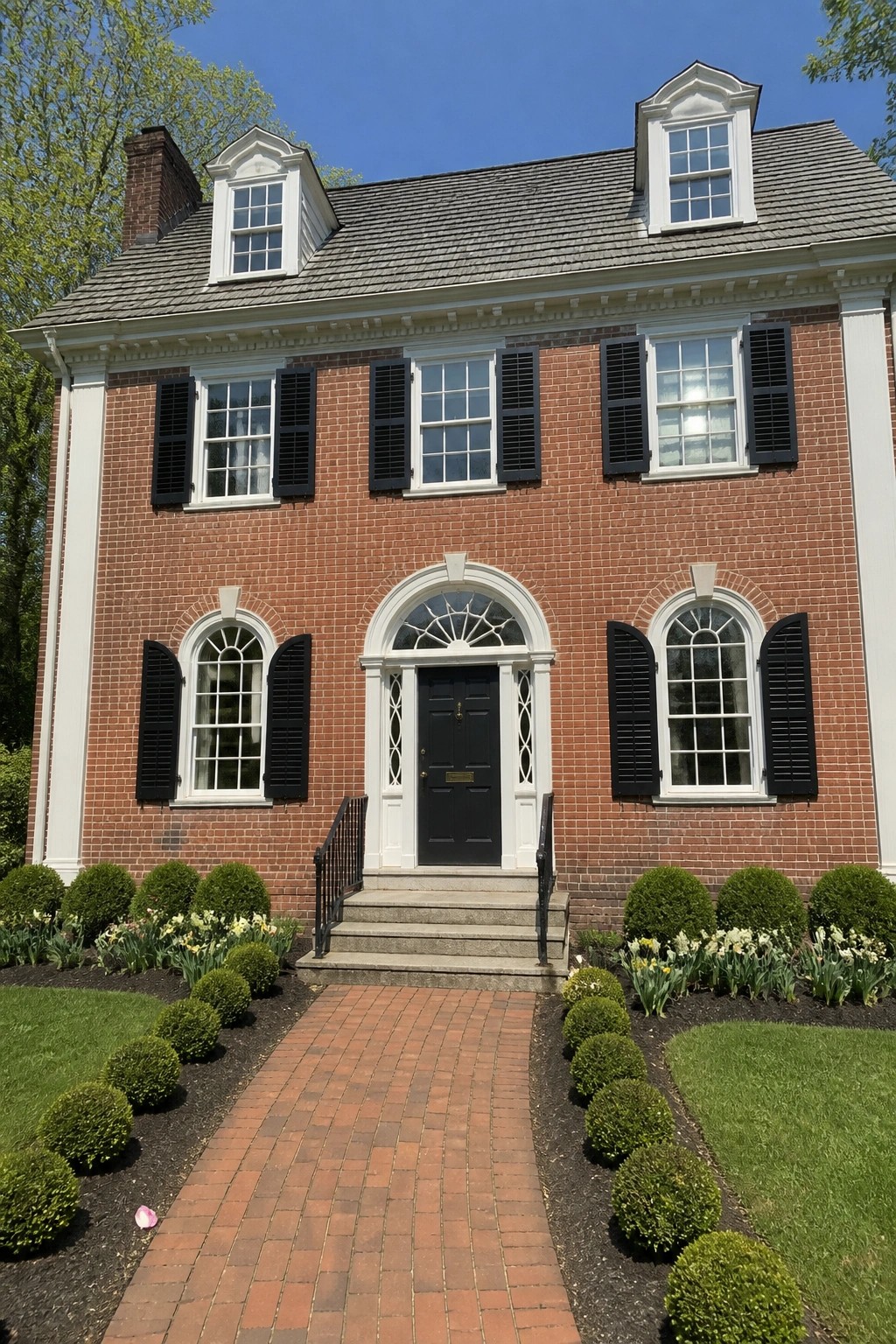 A two-story red brick colonial house with a centered black door under an arched transom window, symmetrical windows with black shutters on both floors, and two dormers on the roof.