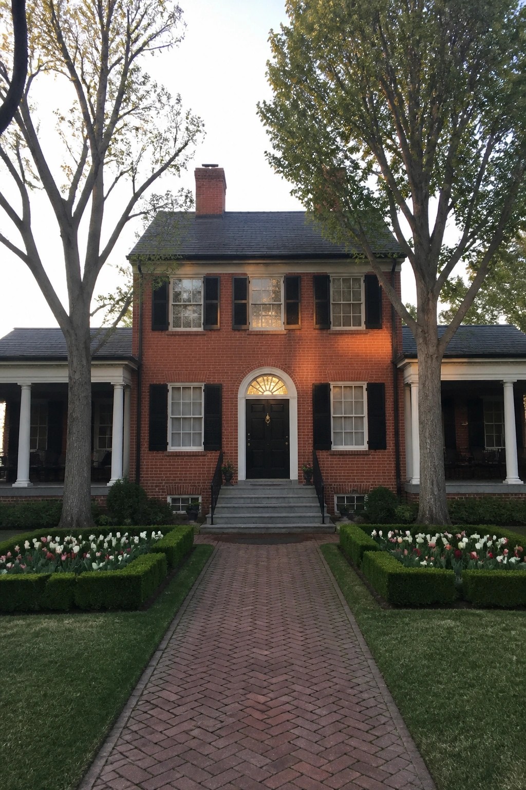 Red brick colonial house with black shutters, brick path, and tulip gardens