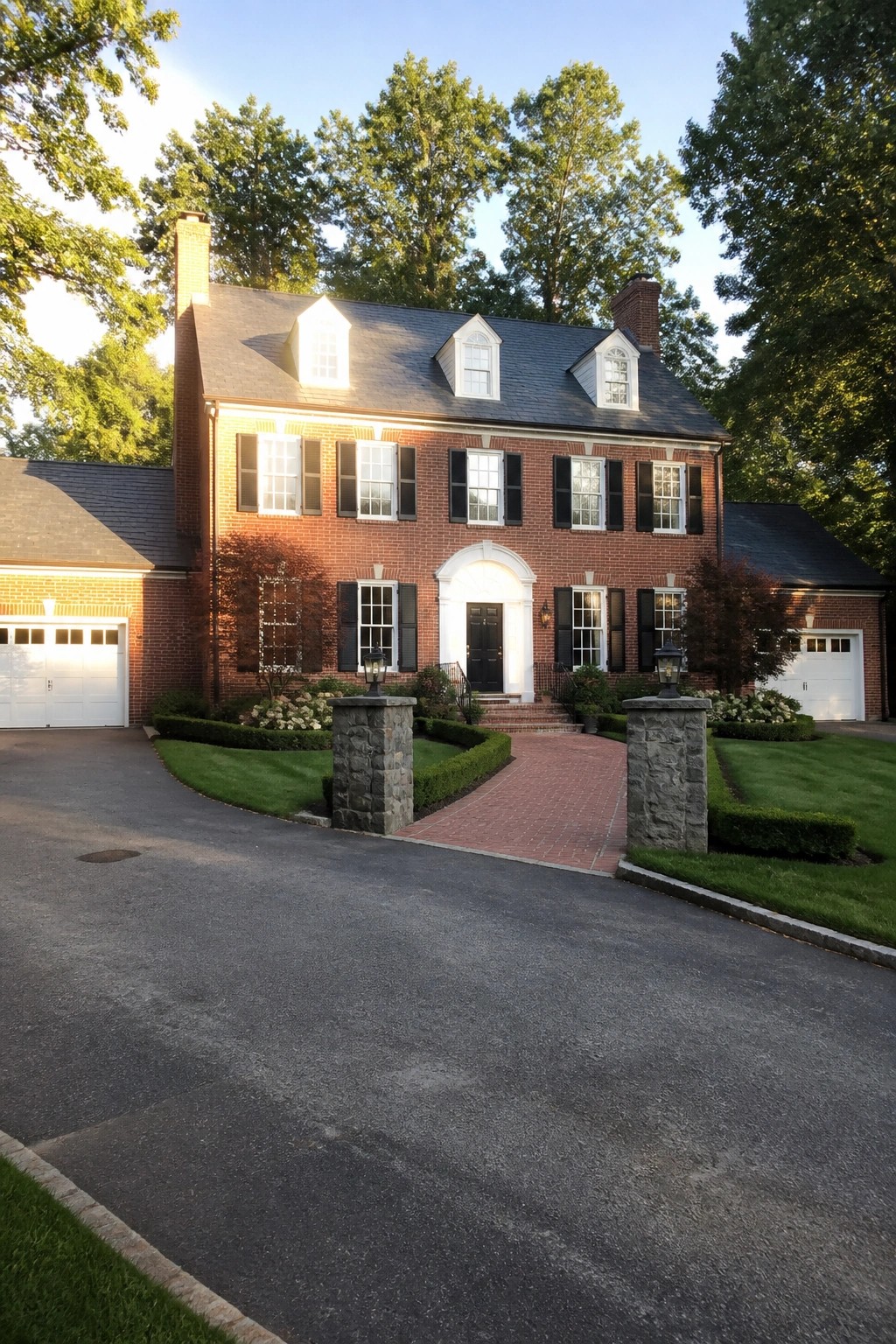 A red brick colonial house viewed from the front drive, showing a centered black door under a white arch, evenly spaced windows with black shutters, and matching side wings with garages.