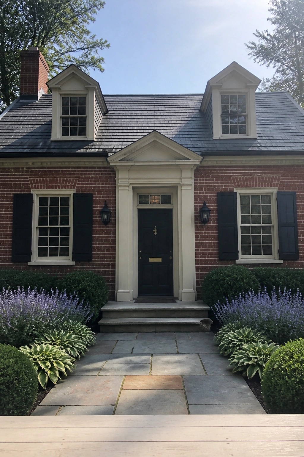 A symmetrical red brick colonial house exterior with a centered dark door under a white pediment, flanked by windows with black shutters and two dormers on the roof.