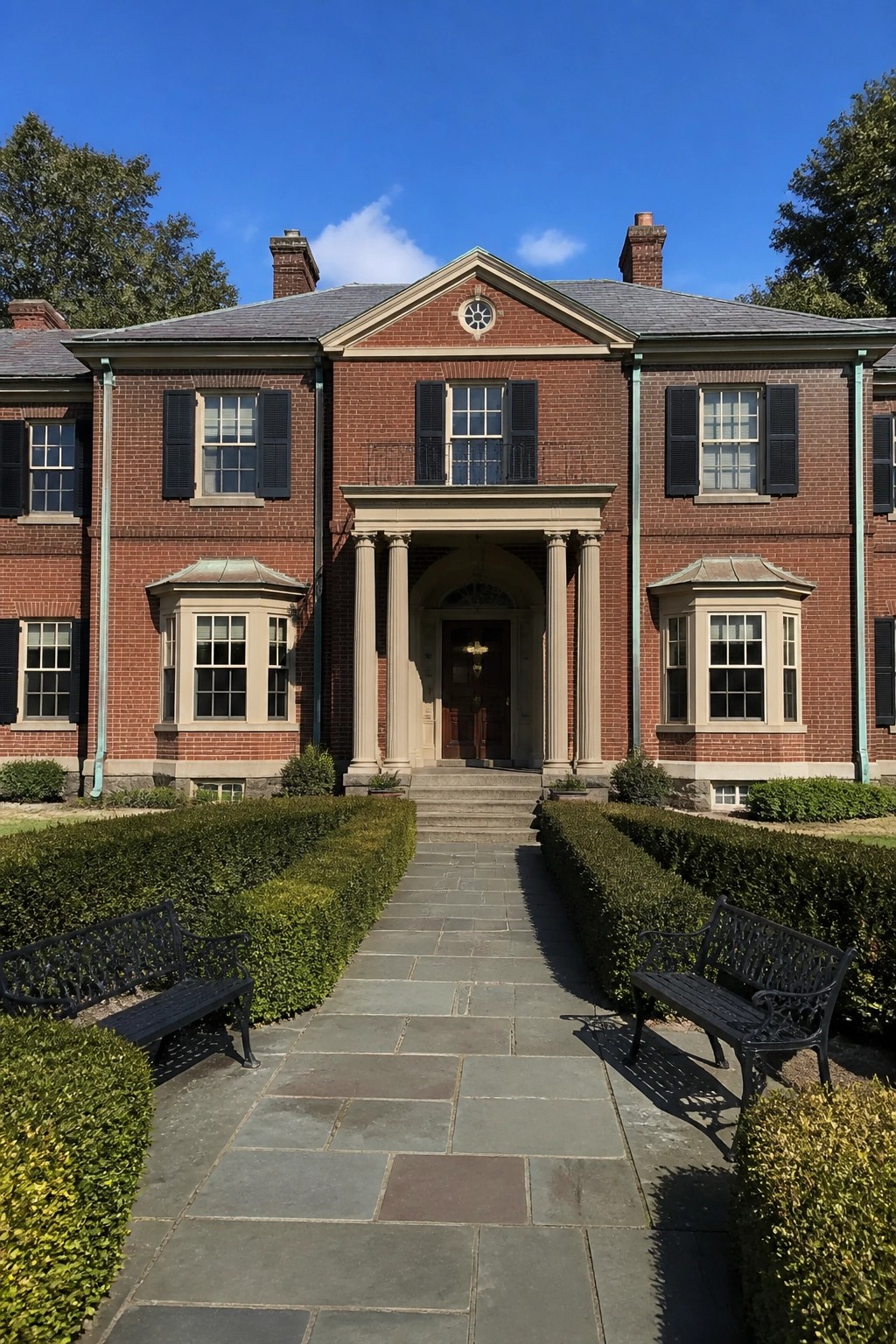 Red brick mansion with columned portico, stone path, black benches, and hedges