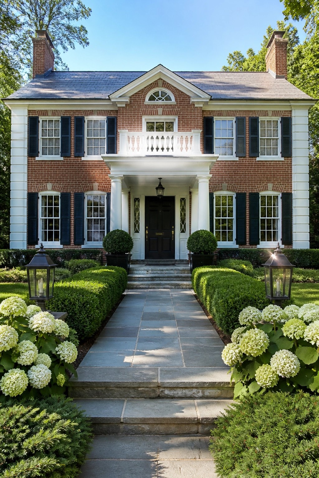 A two-story red brick colonial house with white trim, black shutters, a central portico over the front door, and a stone pathway lined with shrubs and white flowers.