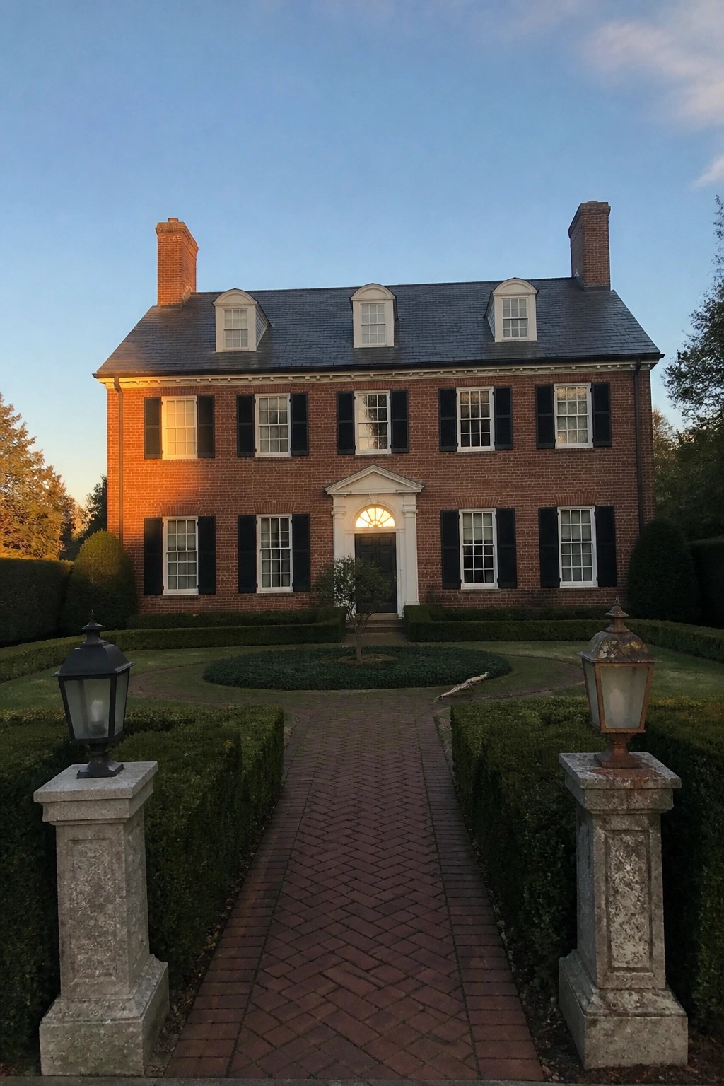 A red brick colonial house with a central white portico entrance, evenly spaced windows with black shutters, two brick chimneys, and a brick pathway leading to the door.