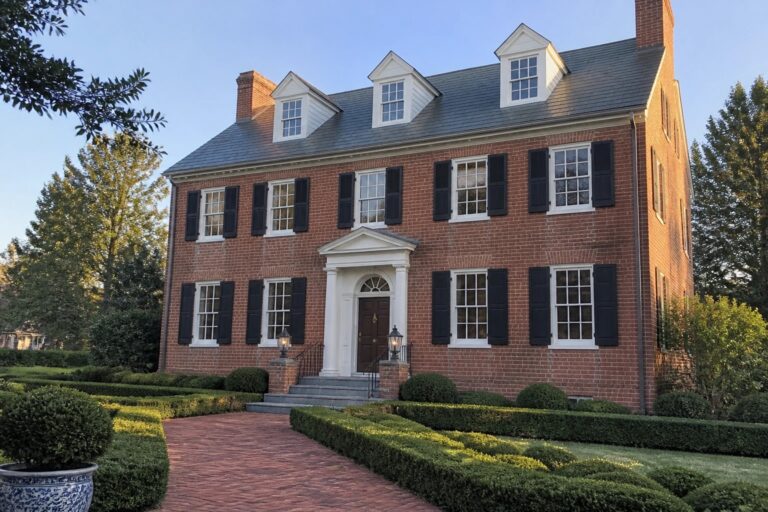 Red brick colonial house with black shutters, white portico, and manicured boxwood hedges