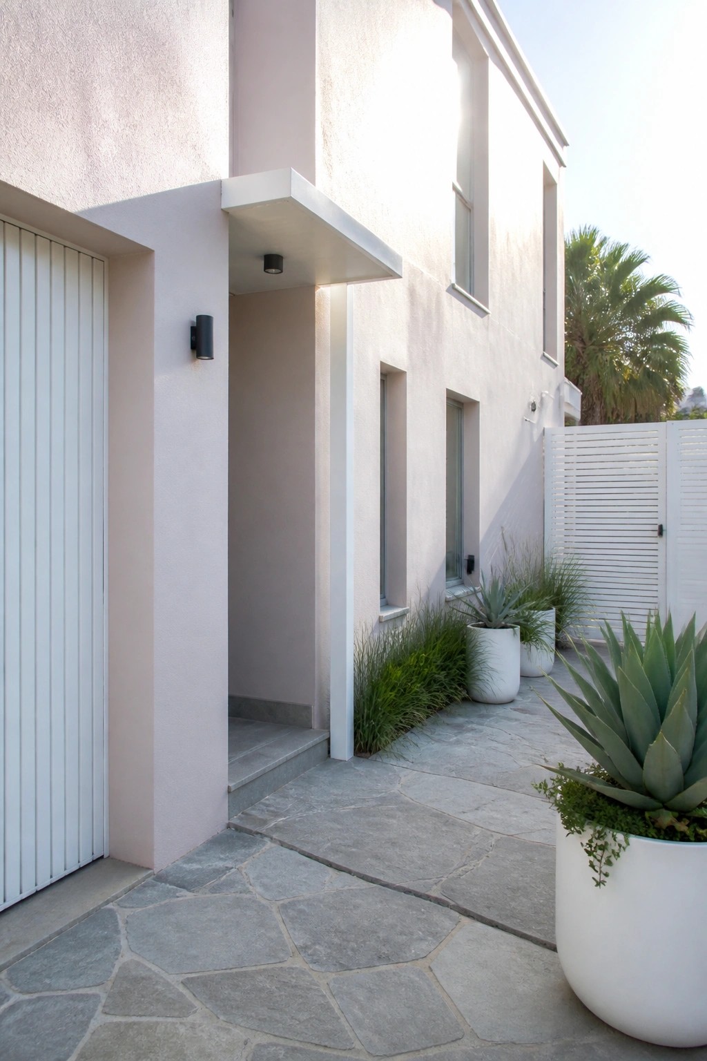 A modern two-story house with soft pink stucco walls, white trim, a recessed entry, and a stone pathway lined with potted plants.