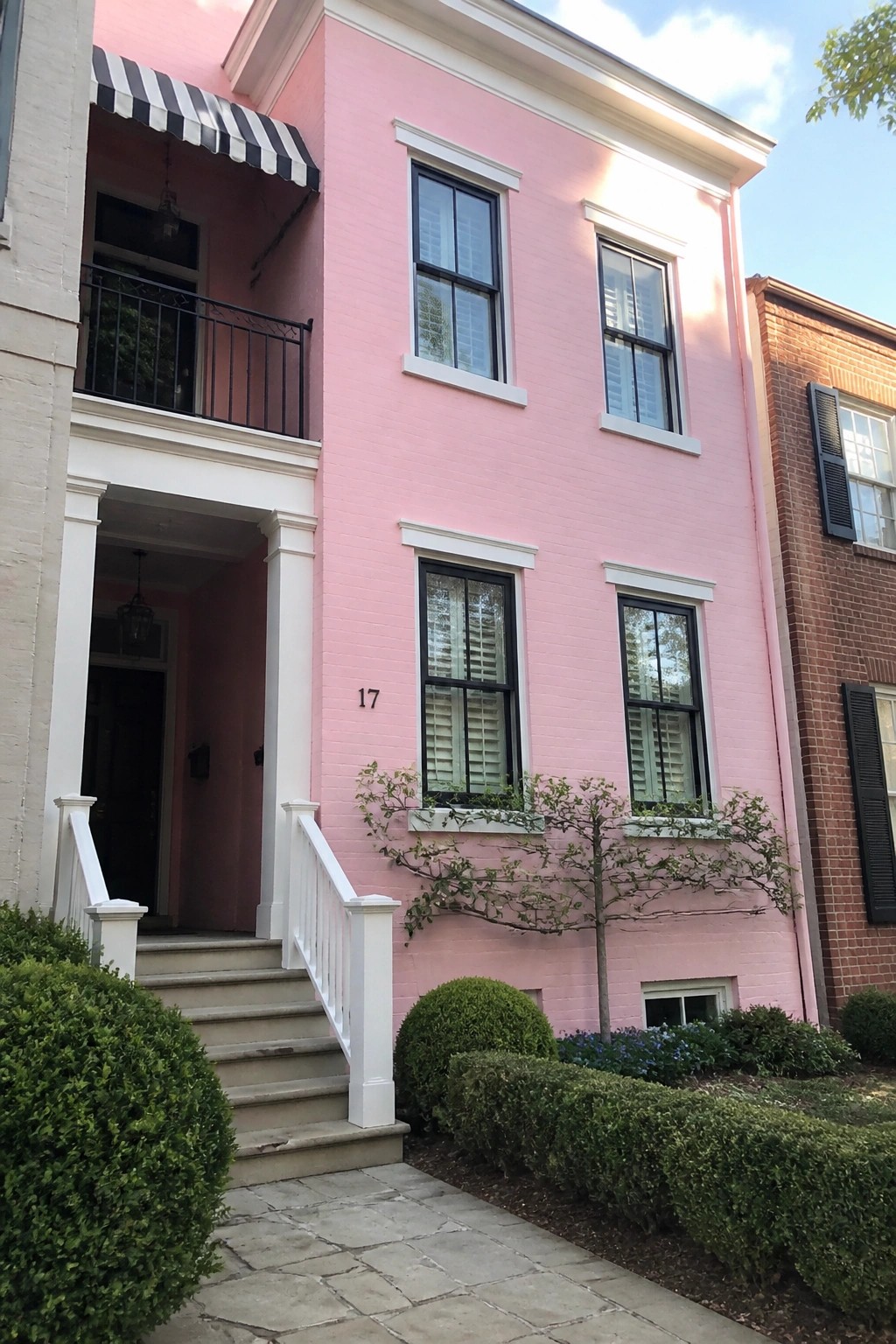Pink brick townhouse with white trim, black windows, striped awning, and garden.
