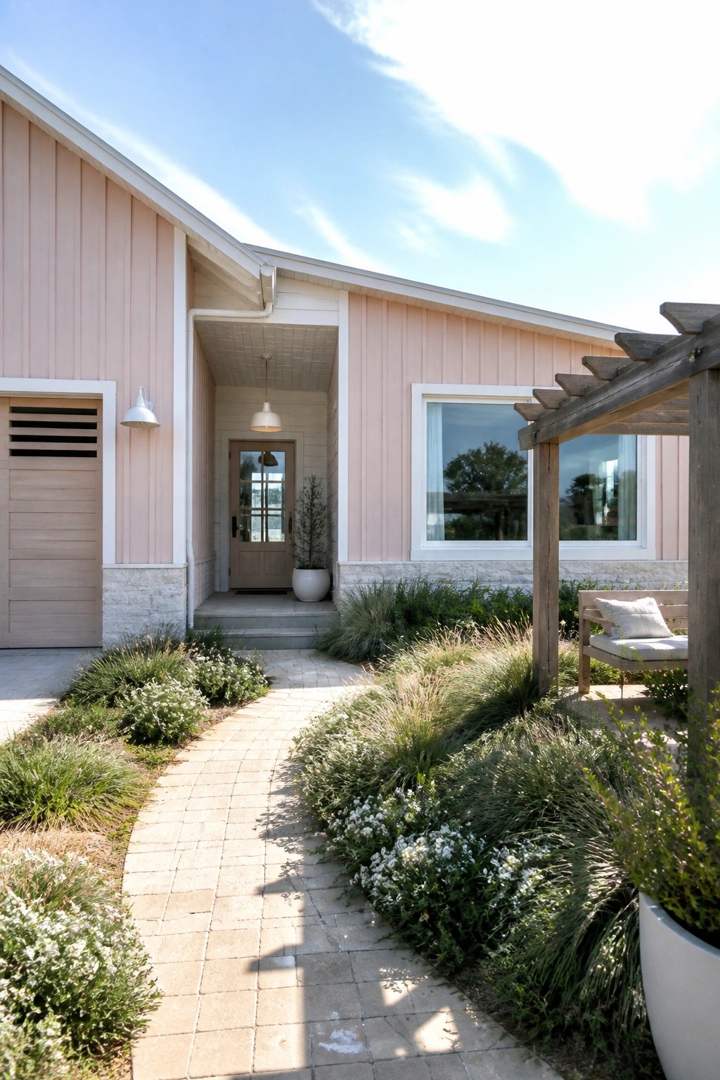 A single-story house with light pink vertical siding, white trim, and a stone base has a paved path leading to a front door, with garden beds along the walkway and a wooden pergola on the right.