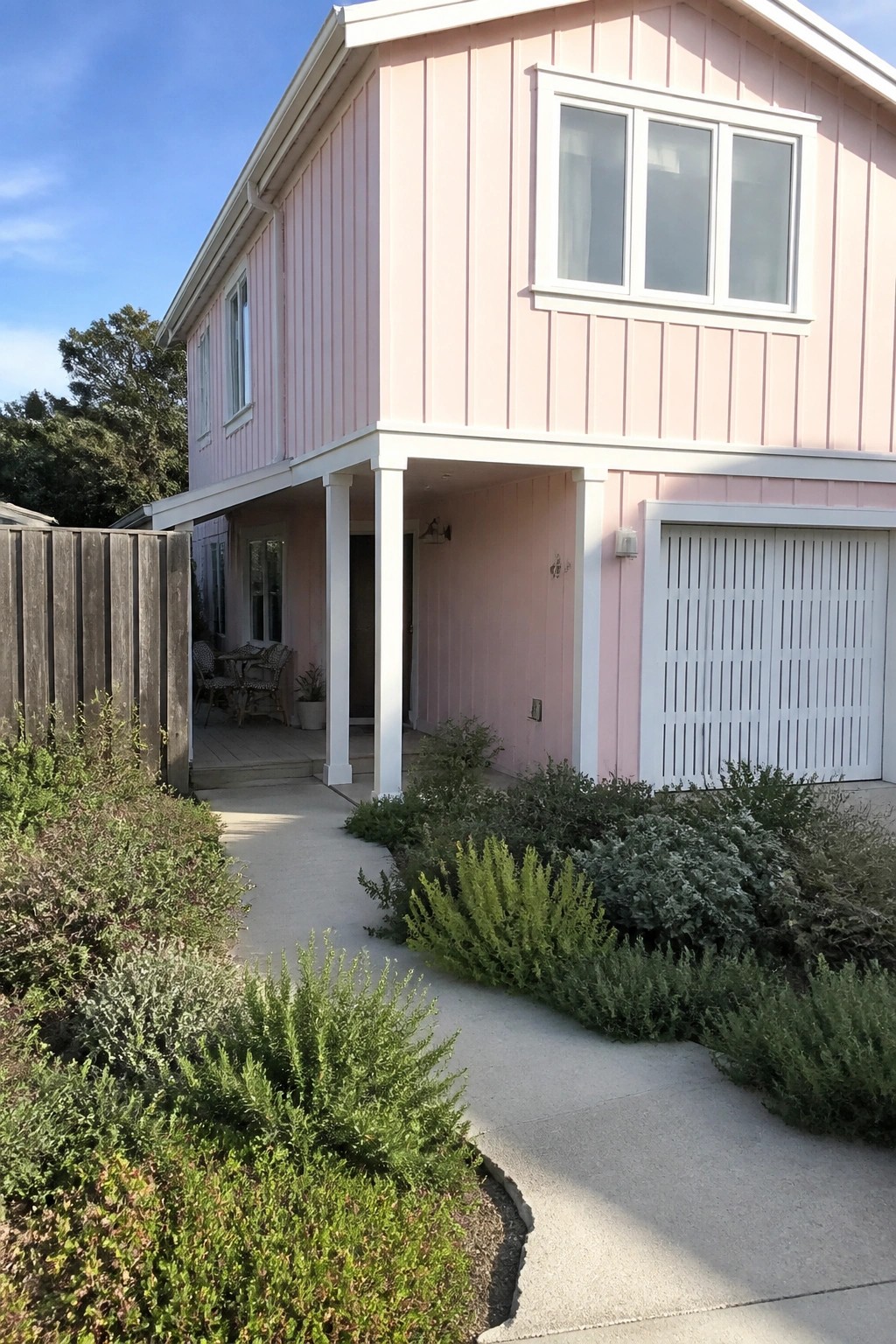 A two-story pink house with white trim, a white garage door, and a concrete walkway lined with green shrubs.