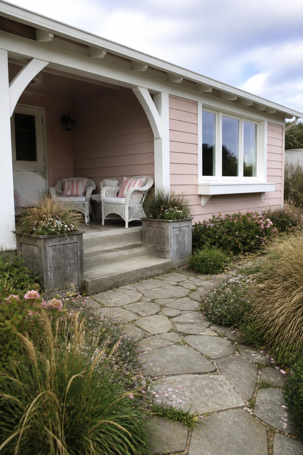 A single-story house with light pink siding and white trim, featuring a covered porch with wicker seating and a flagstone path leading through garden beds.