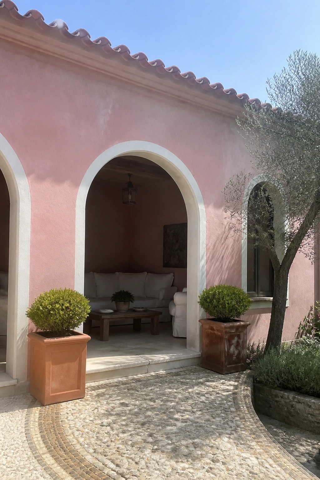 A pink stucco house with white arched openings, two terracotta planters, and a cobblestone courtyard.