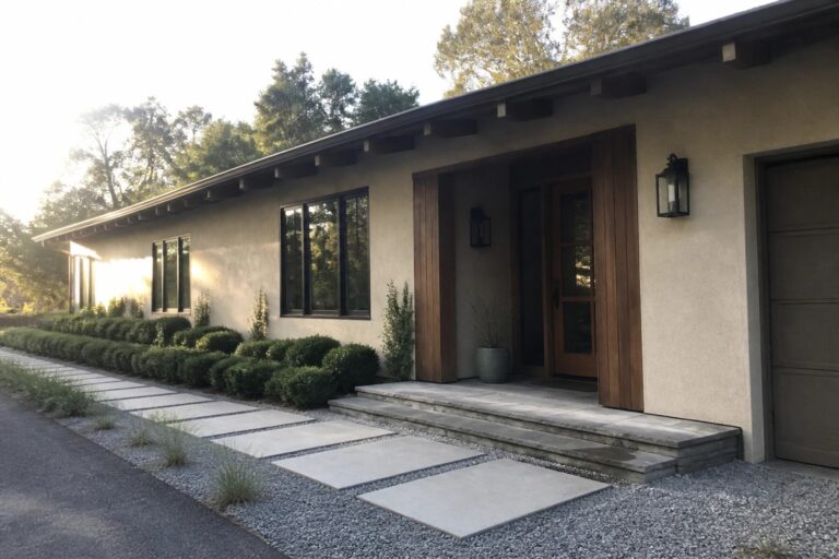 Beige ranch house exterior featuring vertical wood siding around the front door, black-framed windows, a stone porch, and a gravel path with concrete pavers.