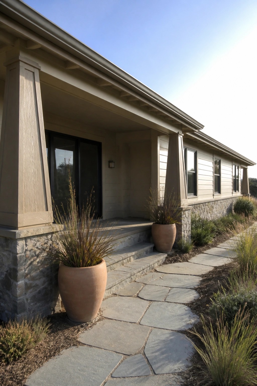 A beige ranch house with a covered porch, columns featuring stone bases, a stone foundation, and a stone pathway along the front.