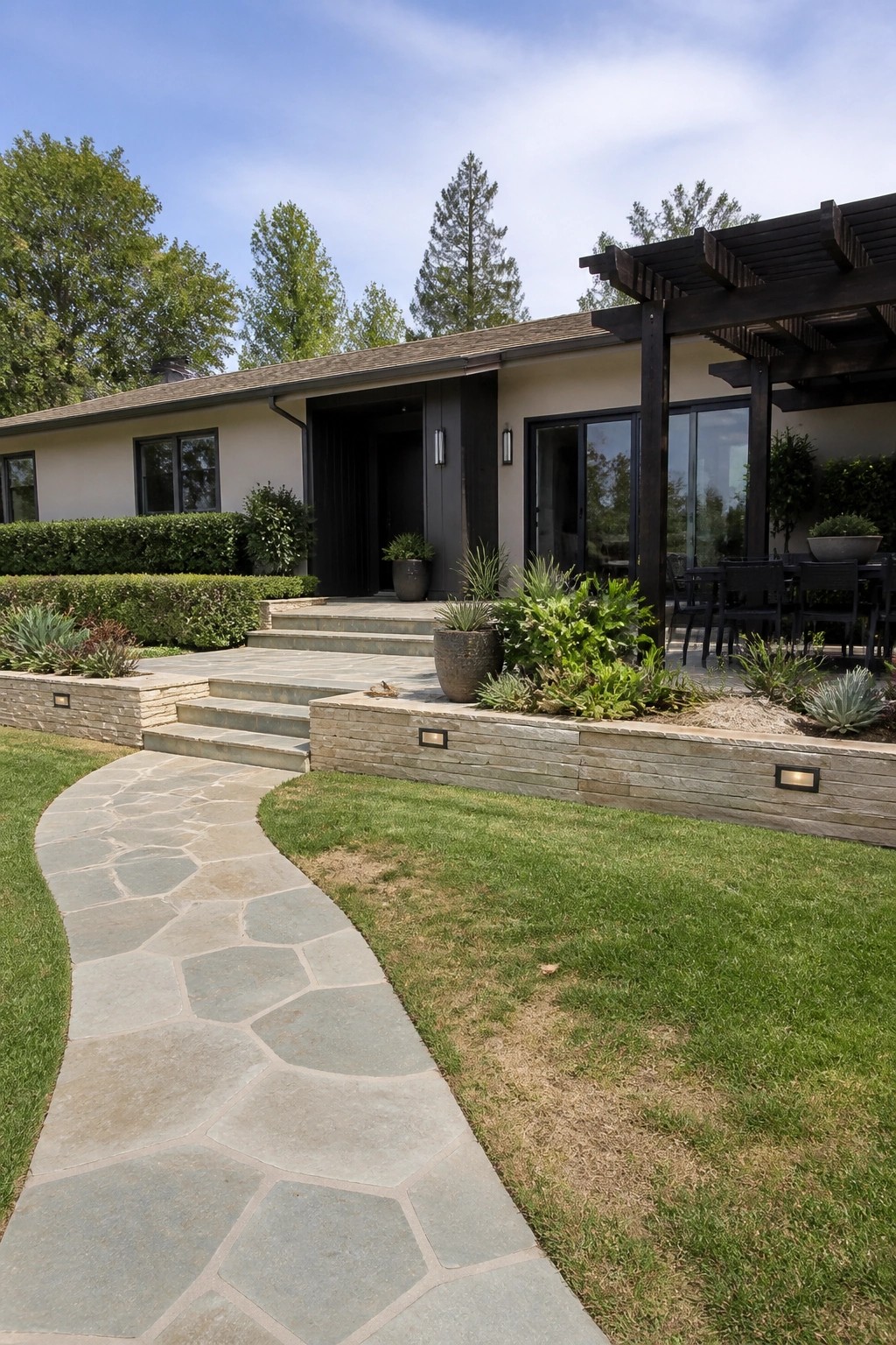A beige ranch house exterior with dark trim, a curved flagstone path, and raised stone retaining walls with plantings along the front.