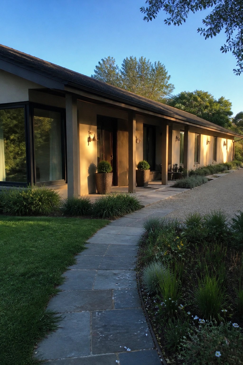 A long beige ranch house exterior with a covered walkway supported by columns, wall sconces, large windows, and a stone path bordered by landscaping.