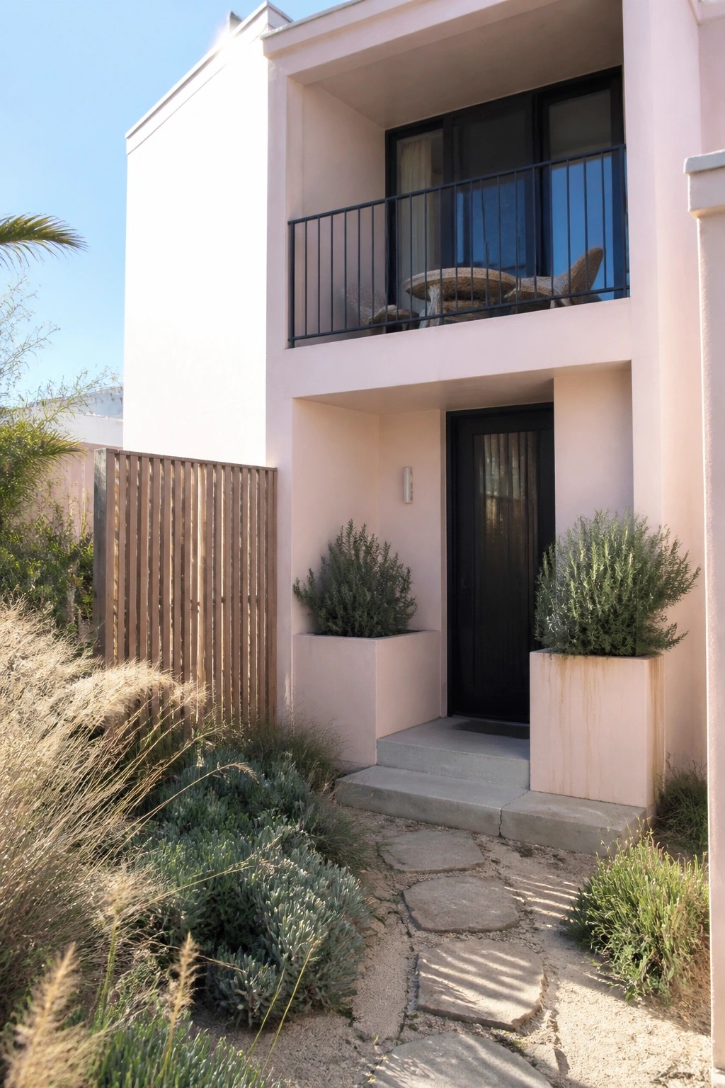 Pink modern villa entrance with black door, balcony, planters, and stone garden path.