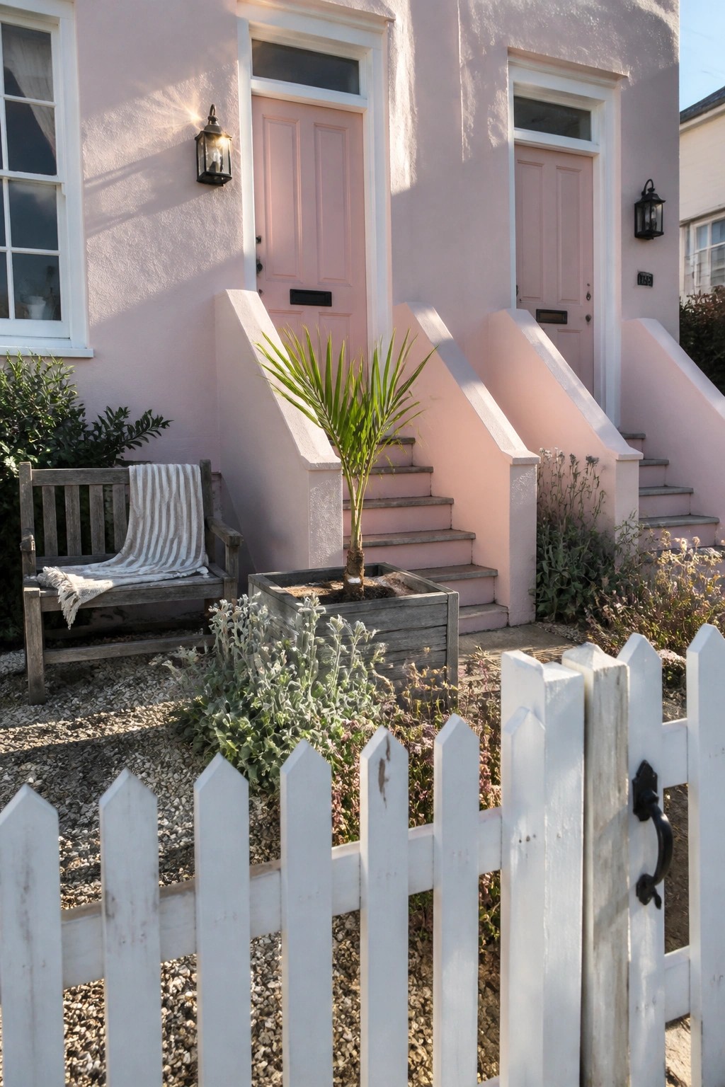 Pink house with steps to doors, wooden bench, potted palm, and white fence.