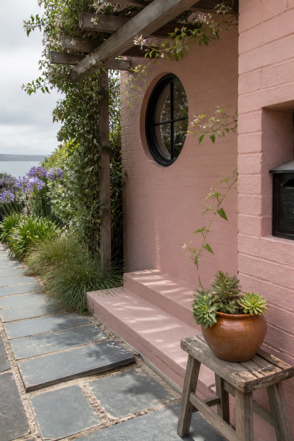 A pink brick house exterior featuring a round black window, wooden pergola overhead, stone pathway, and potted plants on a small wooden stool.