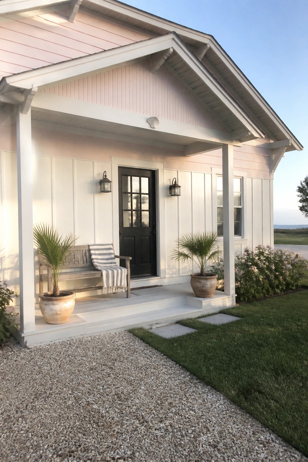 Front view of a light pink house with white trim, a black front door, covered porch, potted plants, and a wooden bench.