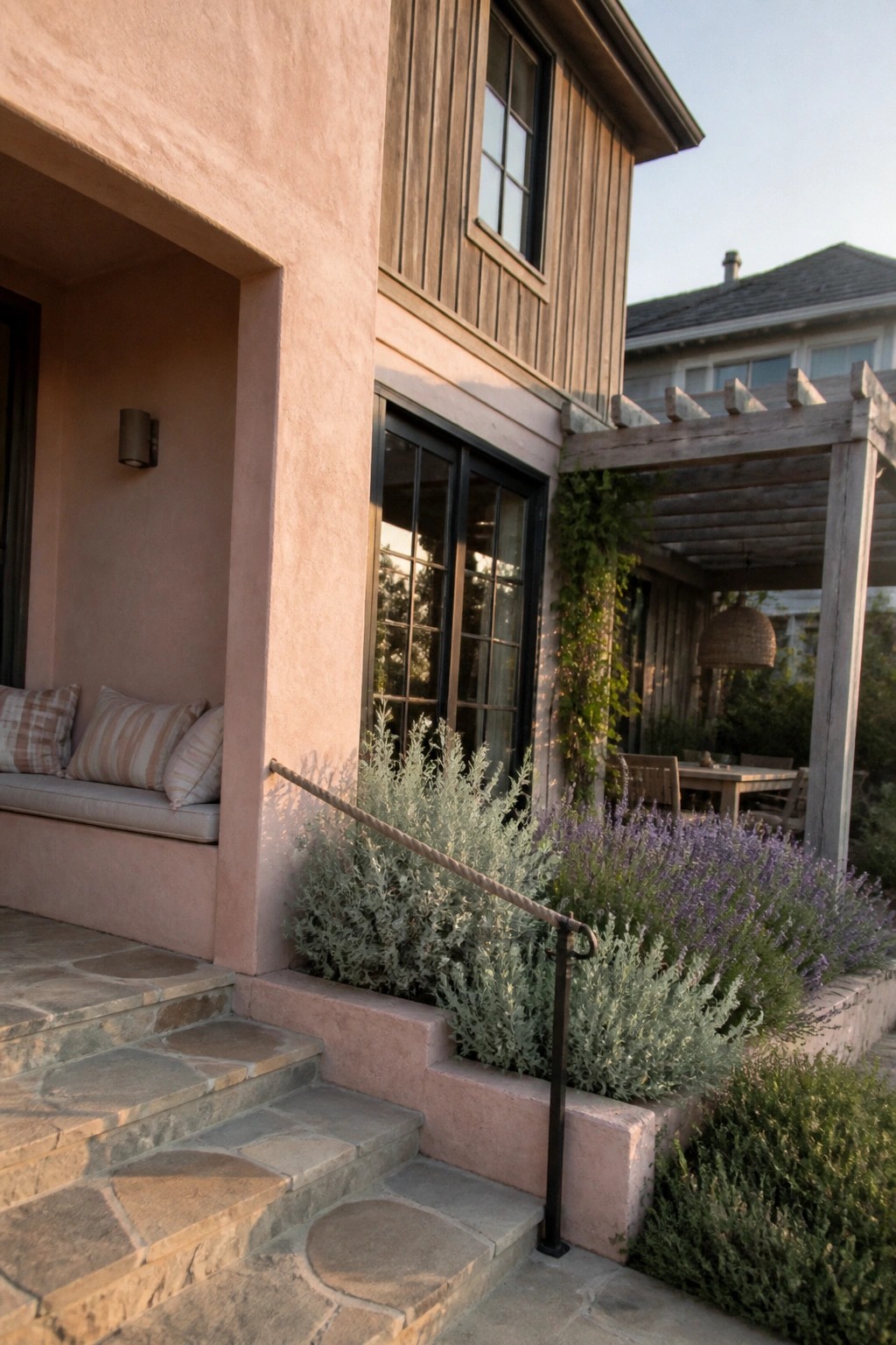 A pink stucco beach house exterior with wooden siding, black-framed windows, stone steps, and purple flowering shrubs beside a wooden pergola.