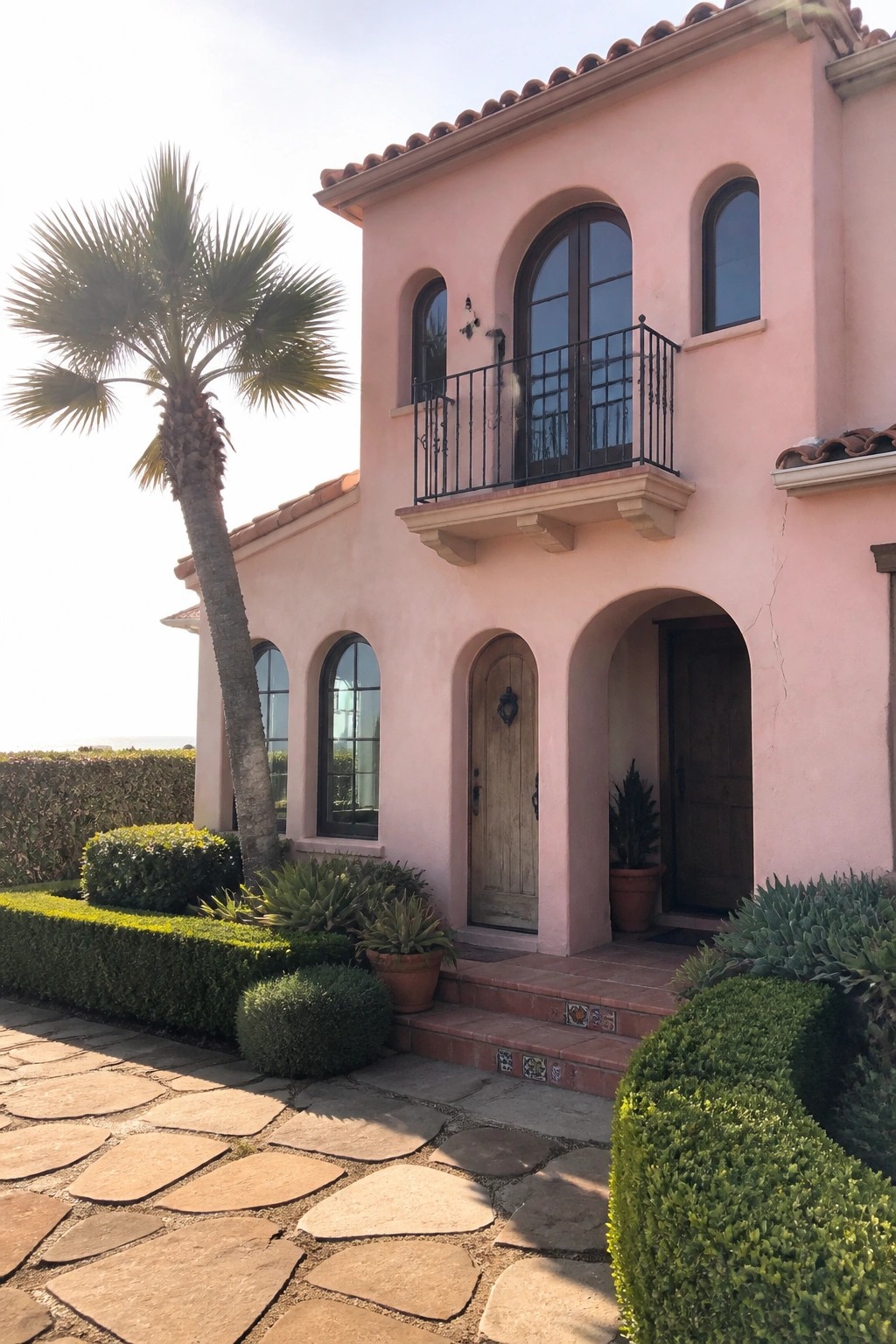 Pink Mediterranean villa with arched windows, balcony, wooden doors, palm tree, and stone path.