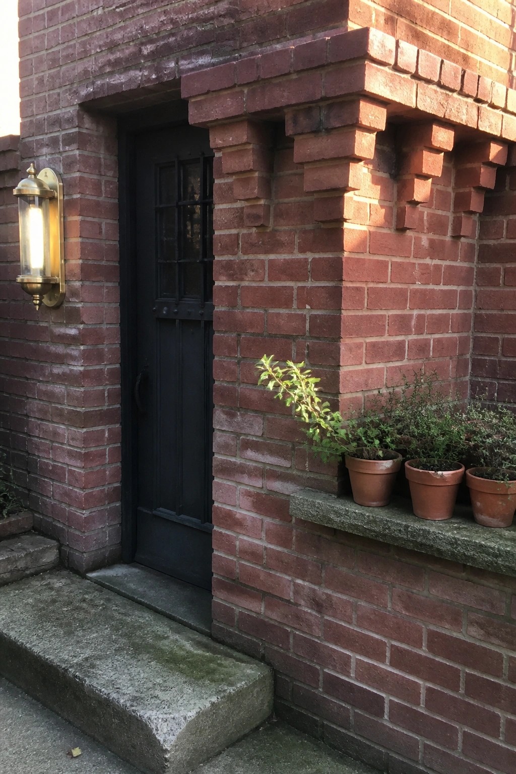A red brick house entrance with a black door, brass wall sconce, and potted plants on a stone ledge.