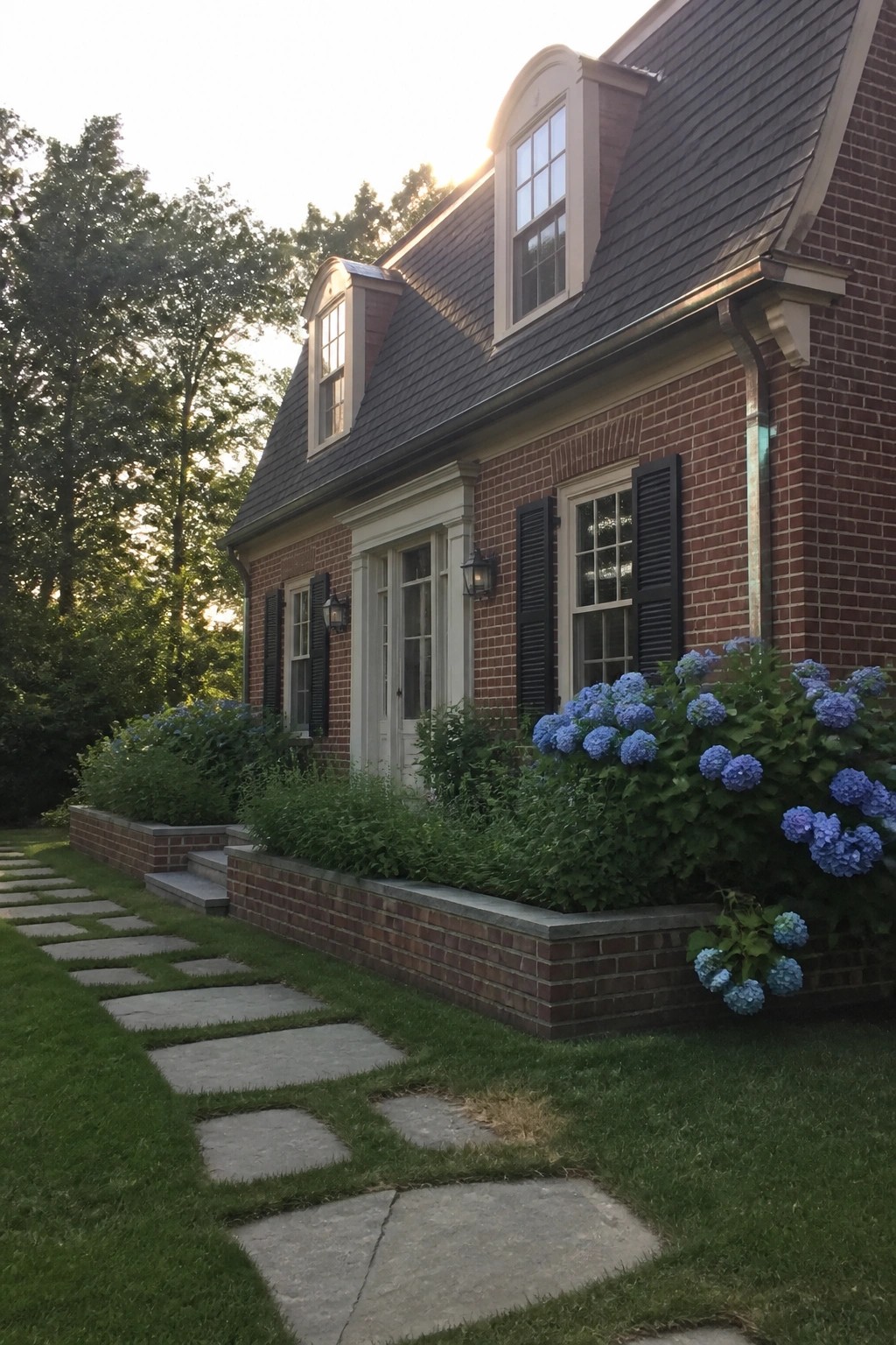 A red brick house exterior with white trim, black shutters, and raised brick planters holding blue hydrangeas along the front walk.