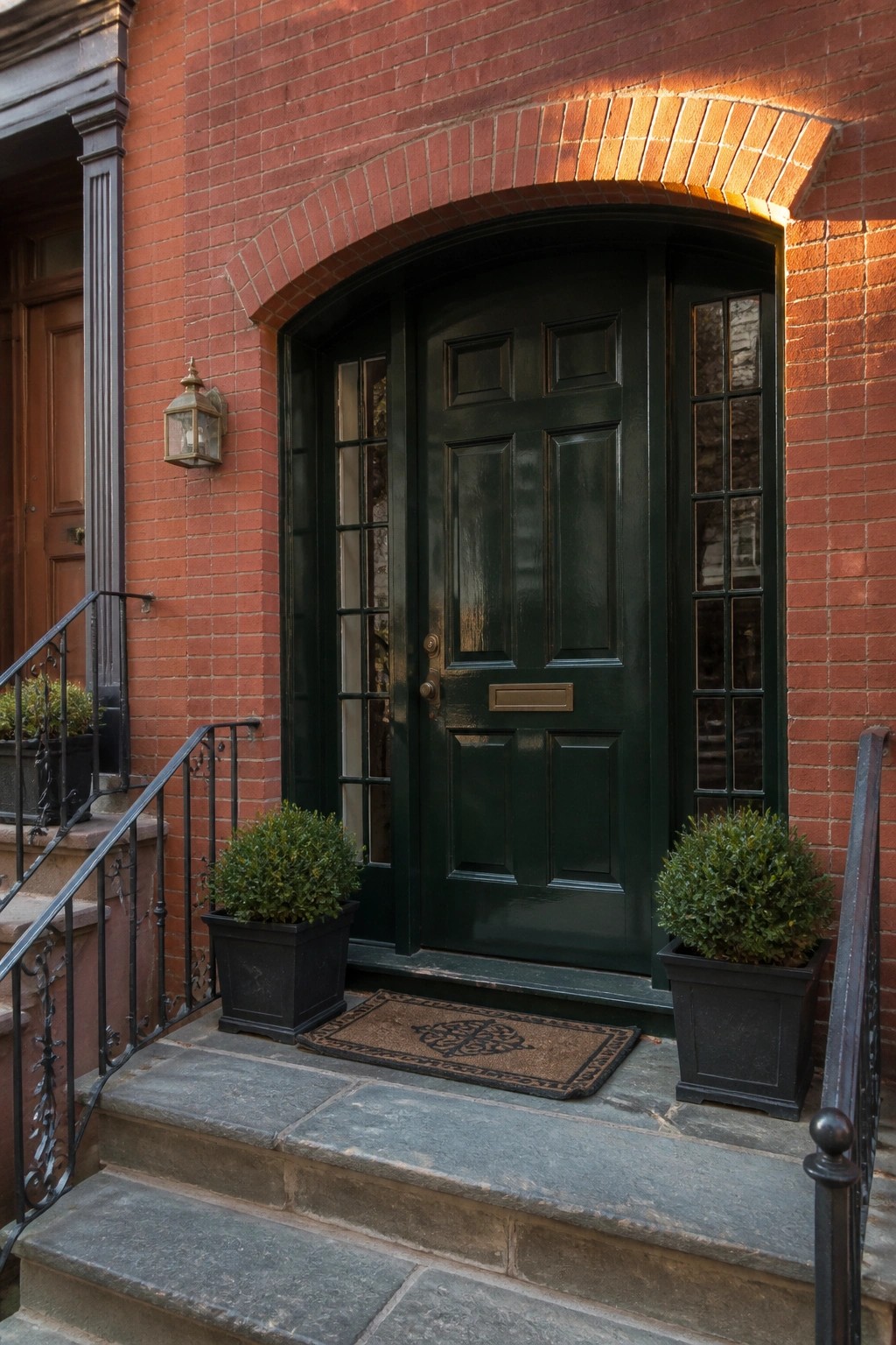 A red brick house with a dark green paneled door centered under a brick arch, flanked by narrow sidelights and stone steps holding two potted shrubs.
