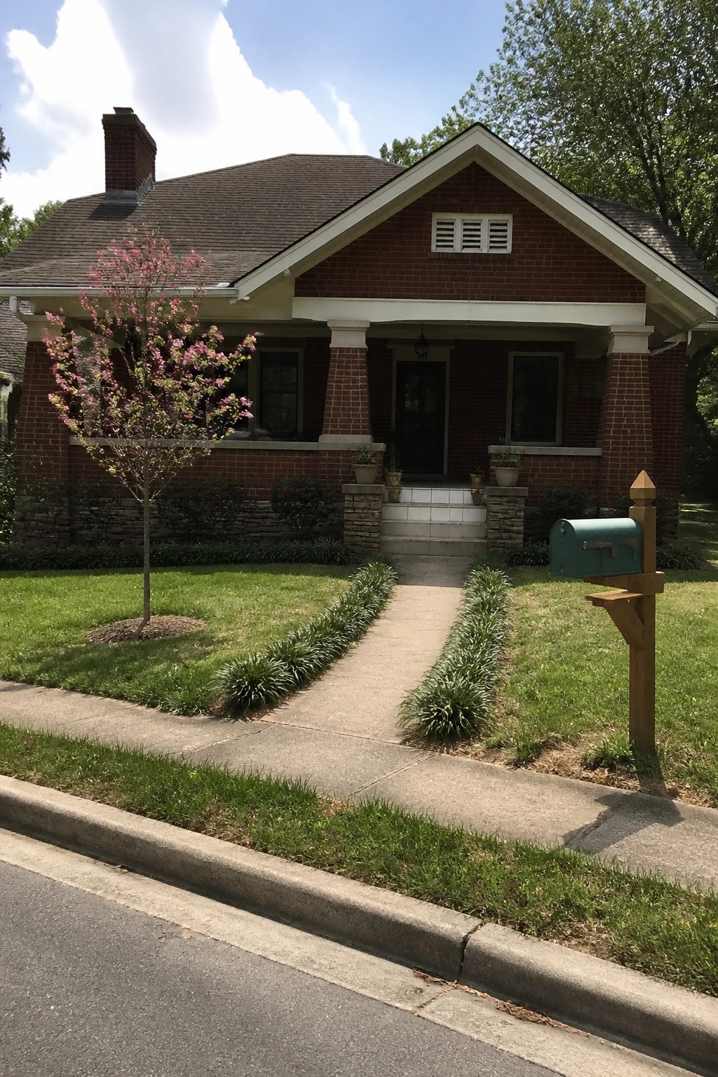Red brick house with porch, pink blooming tree, green lawn, and mailbox by sidewalk
