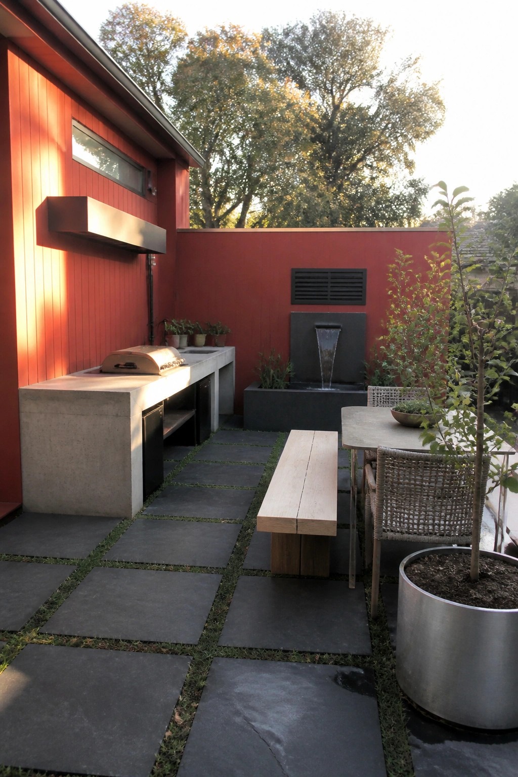 A red house exterior with a concrete outdoor kitchen counter, wooden bench, stone pavers, water fountain, and a small dining table with woven chairs on a patio.