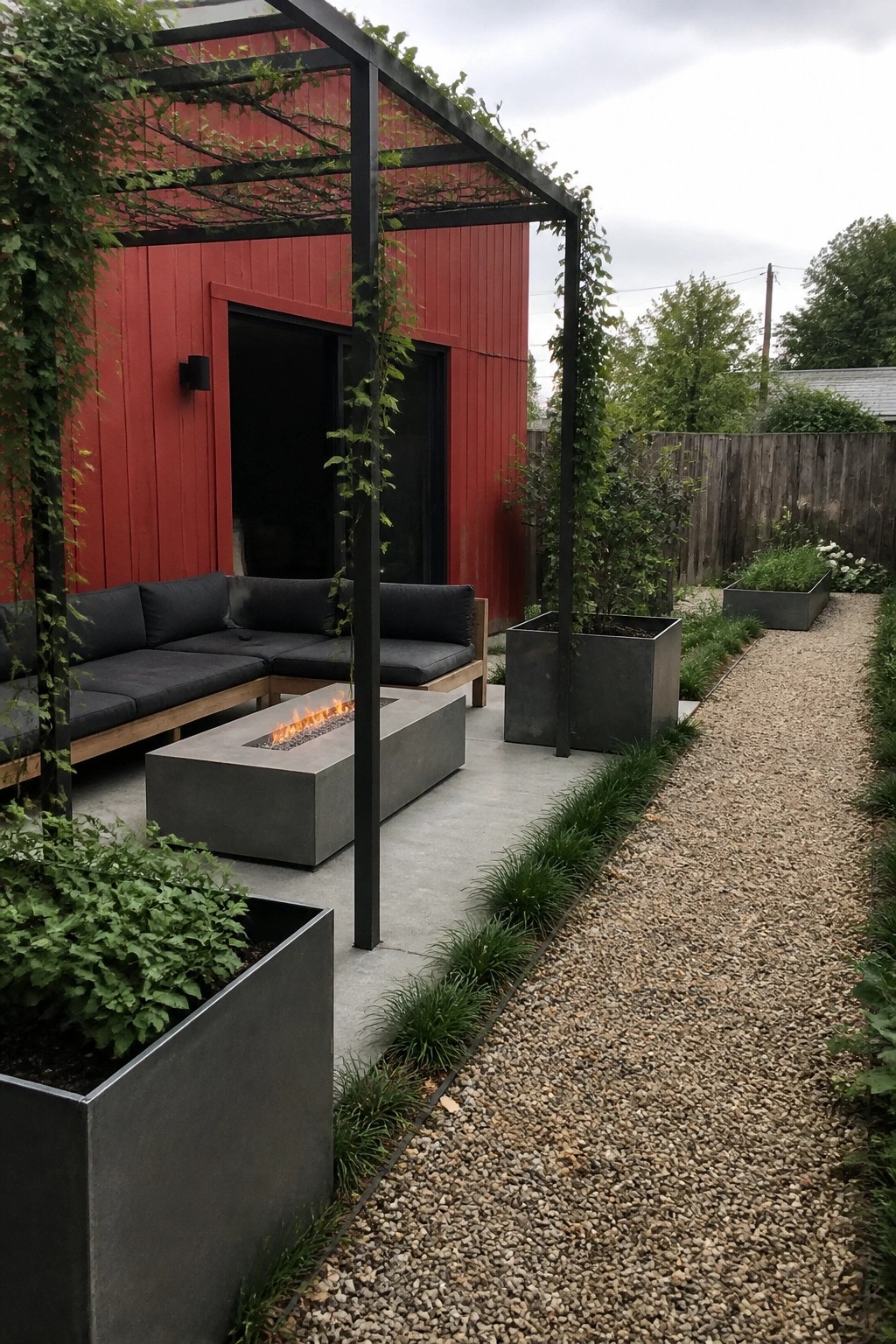 A red house exterior with a black metal pergola over a concrete patio that holds an L-shaped outdoor sofa and a rectangular fire pit, bordered by a gravel path and metal planters.