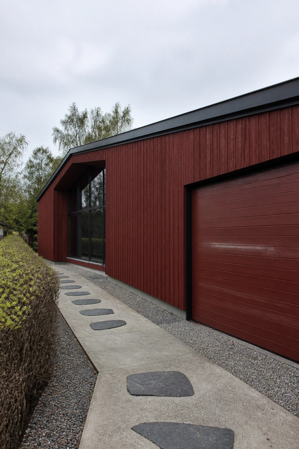 A modern house exterior with vertical red wood siding, a matching red garage door, black roof trim, a large window, and a concrete path with dark stepping stones beside a hedge.