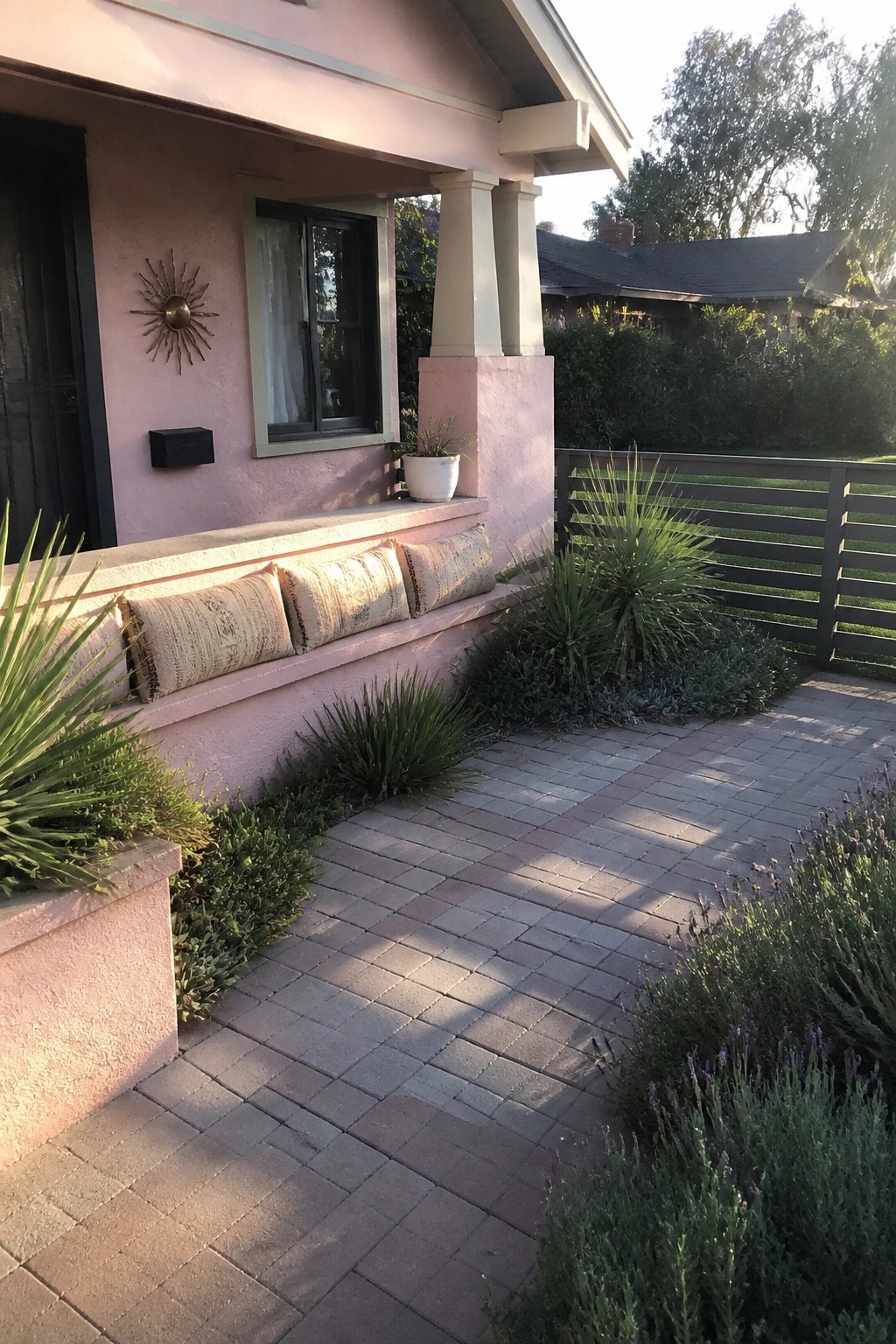 A pink stucco bungalow exterior with a long built-in bench holding patterned cushions beneath a window, surrounded by plants and a brick path.