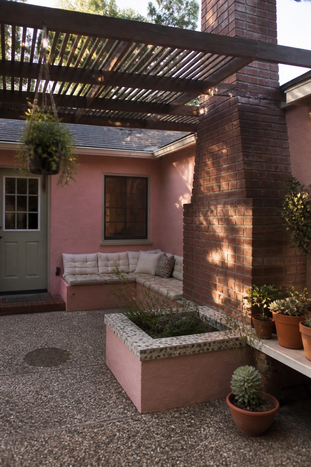 A pink stucco patio features an L-shaped built-in bench with cushions under a wooden pergola, a central tiled planter box, and potted plants beside a brick chimney.