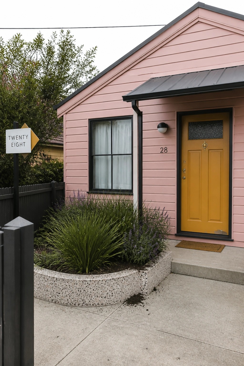 A pink bungalow exterior with a yellow front door, black trim around the door and window, a concrete planter bed with green plants, and a white address sign on a post.