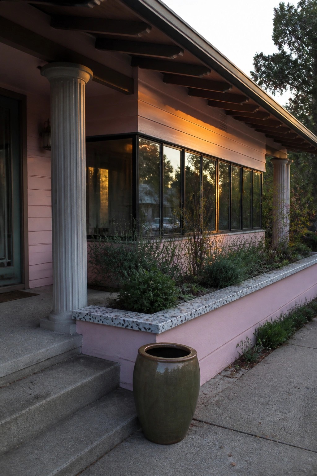 A pink bungalow exterior shows a long window wall, two fluted columns, concrete steps, and a low speckled planter filled with greenery.